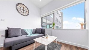 A modern living room with a grey sofa, a white coffee table, and a large window offering a city view. - Captivating 1-bed Apartment 15 min to Londonbridge (Croydon)