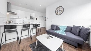 A modern kitchen with white cabinets, a dining area with a white table and chairs, and a grey sofa with a clock on the wall. - Captivating 1-bed Apartment 15 min to Londonbridge (Croydon)