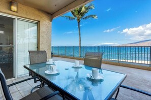 Un balcon avec une table en verre, des chaises et des tasses blanches, donnant sur une plage avec un palmier et un ciel bleu azur.