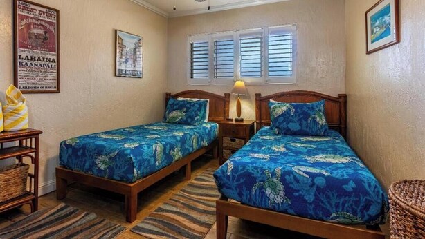A room with two beds, a ceiling fan, a window with shutters, and a wall-mounted lamp.