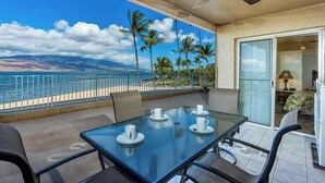 A balcony with a glass table, chairs, and a view of the ocean and palm trees.