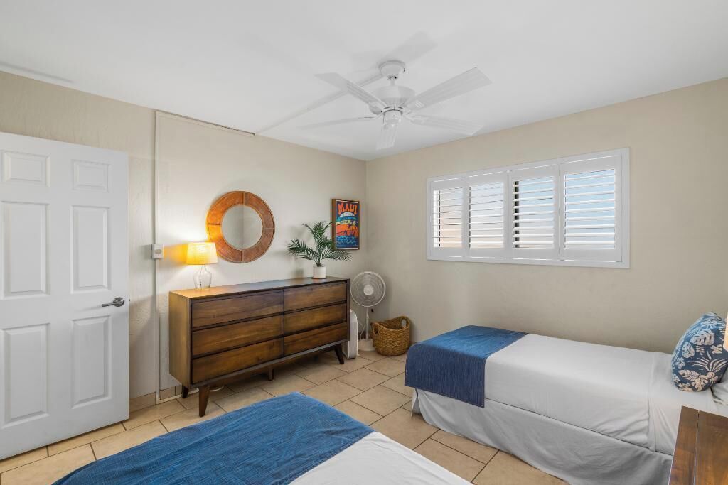 A bedroom with two beds, a ceiling fan, a dresser, a window with blinds, and a door.