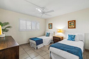 Une chambre d’hôtel avec deux lits, un ventilateur de plafond, une fenêtre avec des stores, une table de chevet avec une lampe et une plante.