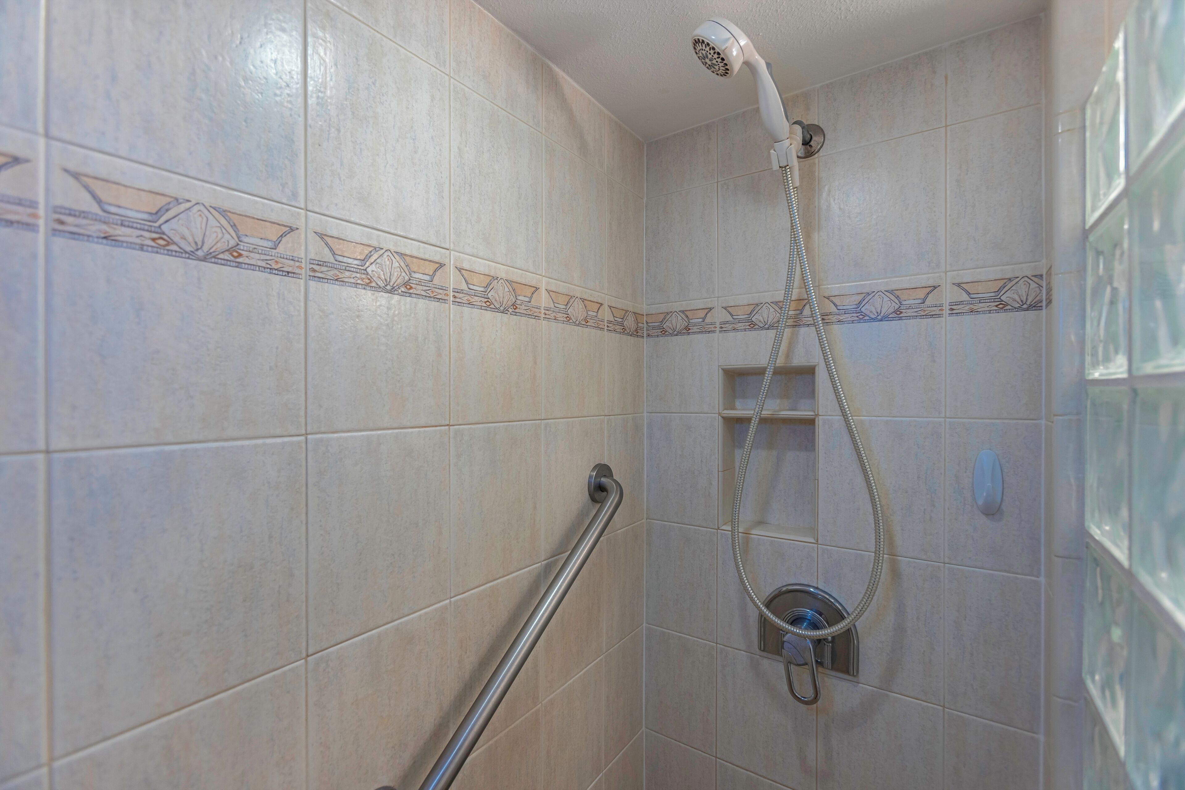 Ducha con cabezal de ducha manual, barra de apoyo y pared de azulejos con detalle decorativo.