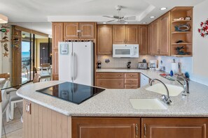 A kitchen with wooden cabinets, a microwave, a refrigerator, a tablet on the counter, and a view of the ocean through a glass door. - Kihei Beach Resort #504- Gorgeous Views and Coastal Elegance (Kihei, Maui)