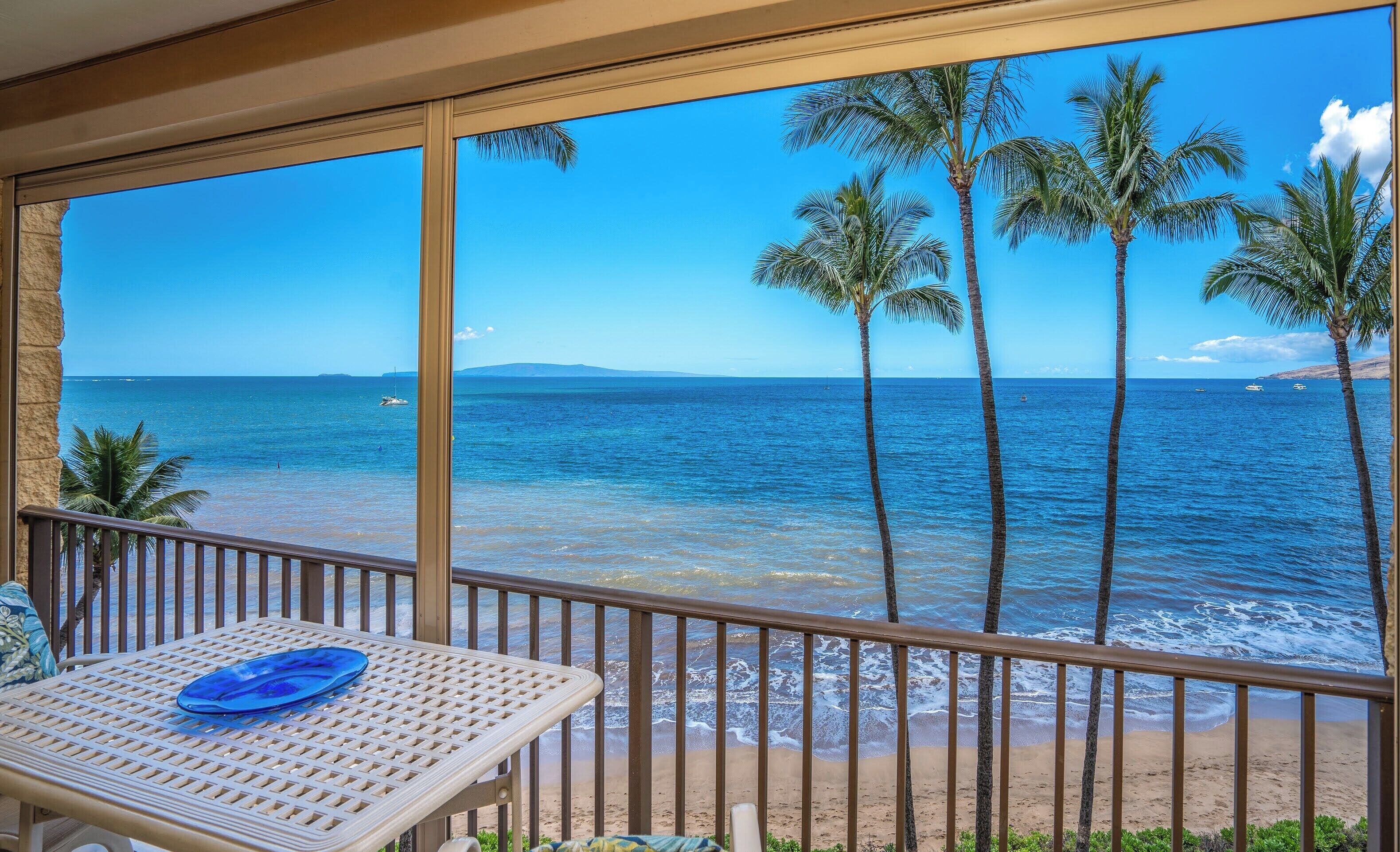 Un balcon avec vue sur l’océan, des palmiers et une table sur laquelle se trouve une assiette bleue.