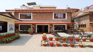 A building with a sign reading "Nepal Cot Hotel Resort." It has a covered outdoor seating area with tables and chairs, surrounded by potted plants. - MeroStay 110 Cottage (Kathmandu)