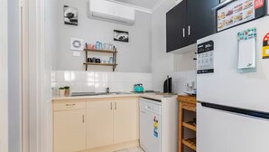 A compact kitchen with white cabinets, a dishwasher, and a refrigerator. There is a small shelf with kitchen items and a few framed pictures on the wall.