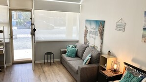 A room with a sofa, a bed, a small table with a lamp, a wall with a poster, and a door leading outside.