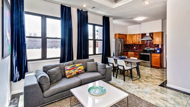 A modern living room with a grey sofa, a colorful cushion, and a white coffee table. A dining area with a table and chairs is visible. The kitchen area includes wooden cabinets and a stove.