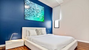 A modern bedroom with a white bed, a nightstand with a lamp, a blue accent wall, and a wooden floor.