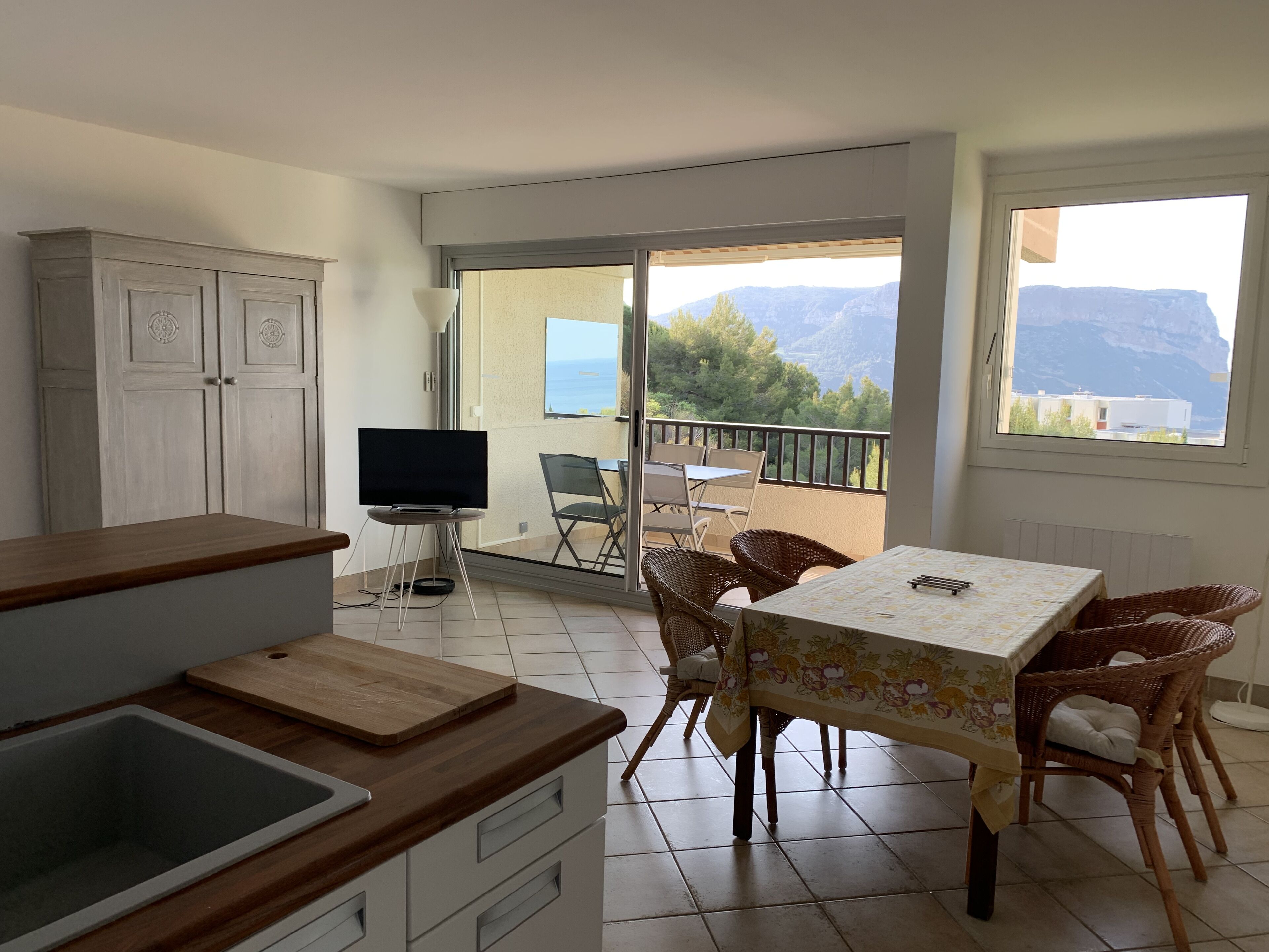 Una cocina con encimera de madera y un lavabo, una zona de comedor con sillas de mimbre y una mesa, y un balcón con vistas a montañas y una mesa con sillas.