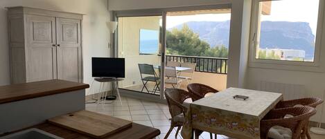 Una cocina con encimera de madera y un lavabo, una zona de comedor con sillas de mimbre y una mesa, y un balcón con vistas a montañas y una mesa con sillas.