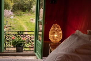 A bedroom with a view of a garden through an open door, a wicker lamp on a bedside table, and a potted plant on the windowsill. - La Maison Odette Chambre D'hotes (aulas)