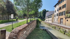 A canal with a brick wall and a grassy area with trees. There is a person riding a bicycle on the path. - Cosy apartment for 6 guests with WIFI, A/C, TV and pets allowed (Lucca)