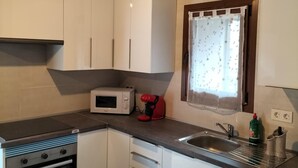 Une cuisine de taille réduite avec des meubles blancs, un four à micro-ondes, un mixeur rouge, un lave-vaisselle et une fenêtre ornée d’un rideau à motifs floraux.