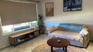 A living room with a sofa, a coffee table, a bookshelf, a potted plant, and a window with a view. - 26 Tambor Drive - Pool and Pet Friendly (Exmouth)
