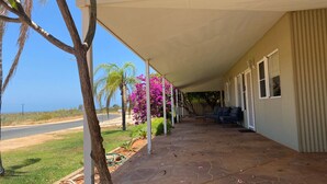 A covered porch with a view of the road and ocean, a patio with a table and chairs, and a garden with flowering plants. - 26 Tambor Drive - Pool and Pet Friendly (Exmouth)