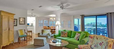 A spacious living room with a green sofa, a floral armchair, and a wooden coffee table. There is a dining area in the background with a chandelier.