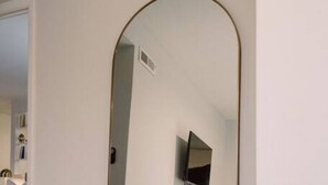 Een kamer met een grote spiegel, een grijze bank en een aan de muur bevestigde televisie.