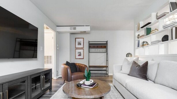 Een moderne woonkamer met een grote flatscreen televisie, een leren fauteuil, een witte bank en een salontafel met boeken en een plant.