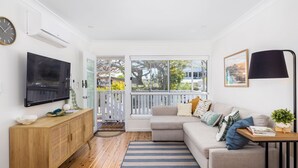 A modern living room with a flat-screen TV, a wooden cabinet, a sofa with cushions, a striped rug, and a large window with a view of trees and houses.