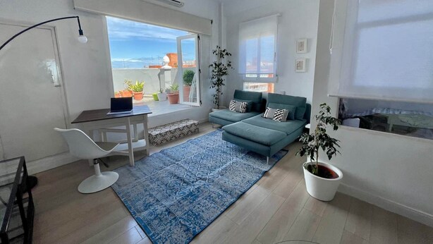 Ein modernes Wohnzimmer mit Sofa, Schreibtisch und Laptop. Es befinden sich Blumentöpfe darin und es gibt einen Blick ins Freie.