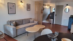 Un salon moderne avec un canapé, une petite table et un coin repas avec une table dressée pour deux personnes. On peut voir des tableaux encadrés au mur, une cloison en bois et, au fond, un espace cuisine.