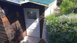 Een klein houten vakantiehuisje met een witte deur, een betegeld terras, en een tuin vol paarse bloemen.