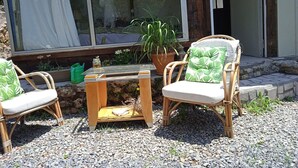 Een terras met rieten stoelen en een kleine tafel, een plant in pot en een raam met gordijn.