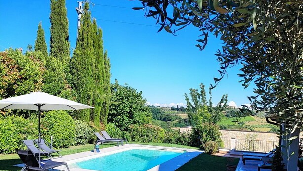 Une piscine entourée de verdure et d’un mur de pierre, avec des chaises longues et un parasol.