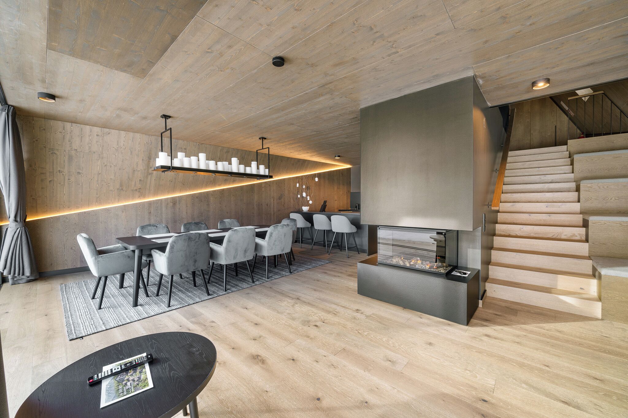 A modern living room with a wooden floor, a fireplace, a dining area with chairs, and a staircase.