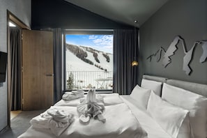 A hotel room with a bed, a view of a snowy mountain, and a table by the window. - TRIFORET alpin.resort by Falkensteiner (Hinterstoder)