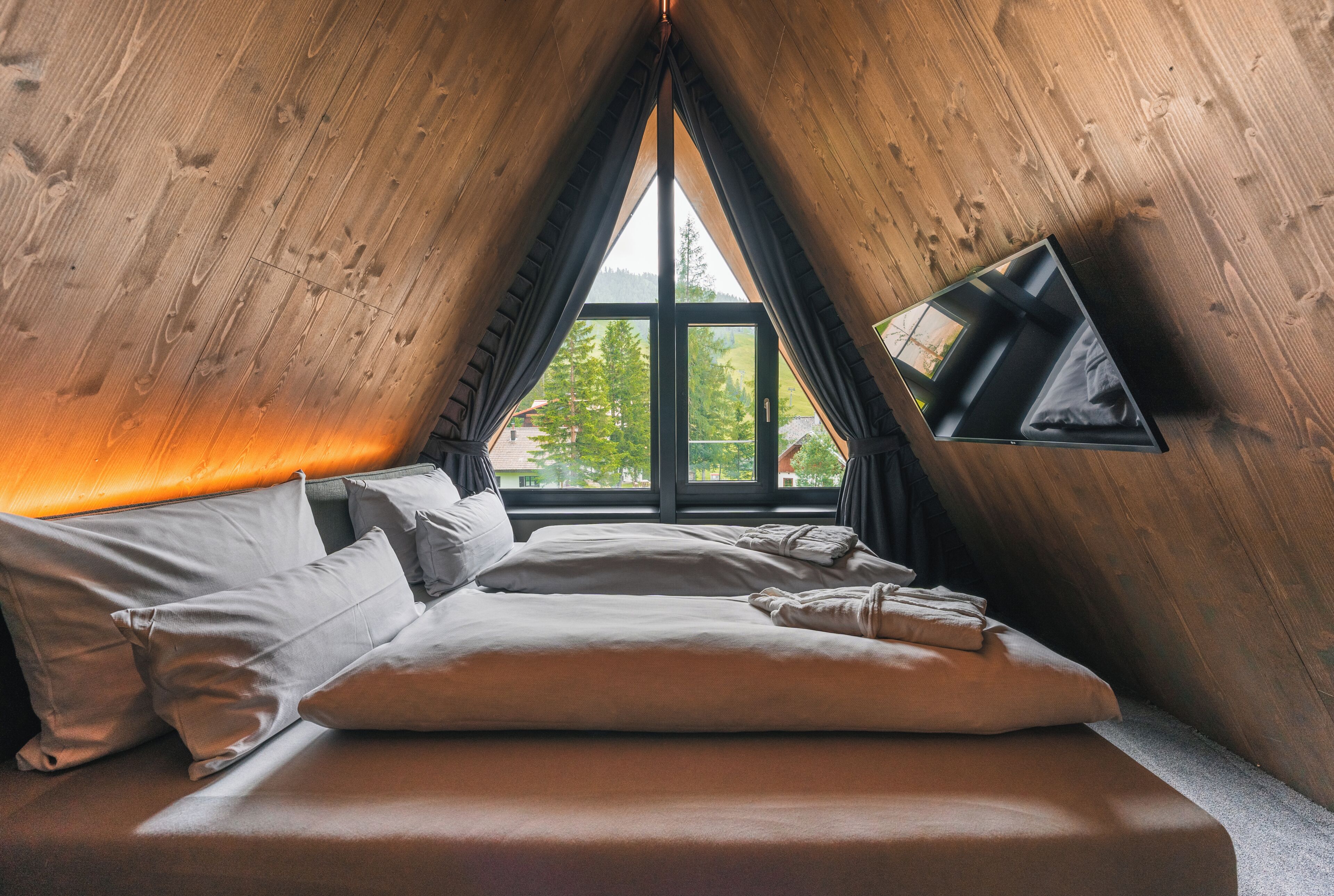 A cozy, triangular bedroom with a large window, a flat-screen TV, and a bed with multiple pillows.