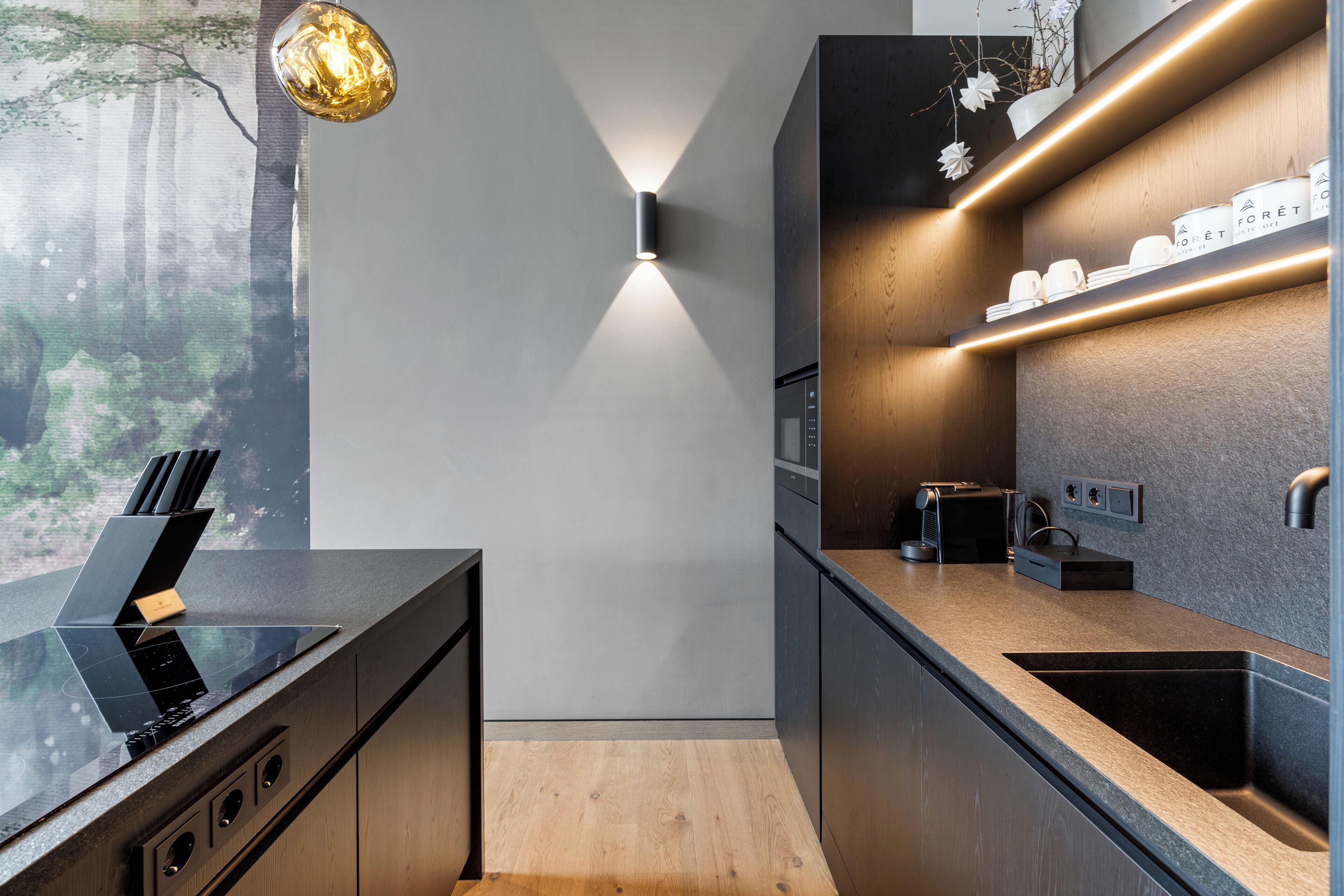 Una cocina moderna con encimera negra, armarios de madera, estufa integrada y una lámpara de pared.