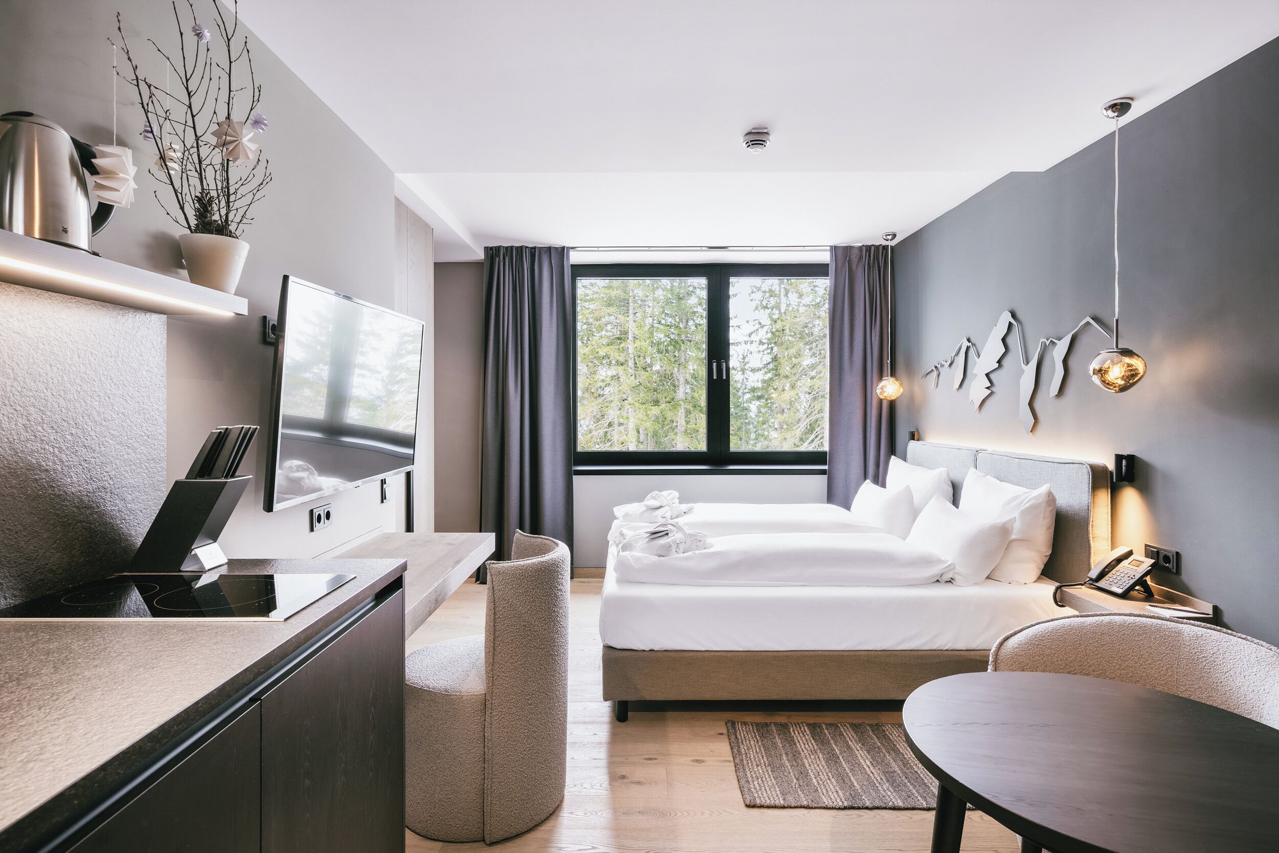 A modern hotel room with a bed, desk, and seating area, featuring a large window and a view of trees.