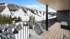 Un balcón con vistas a un paisaje montañoso nevado, suelo de madera y barandilla negra.
