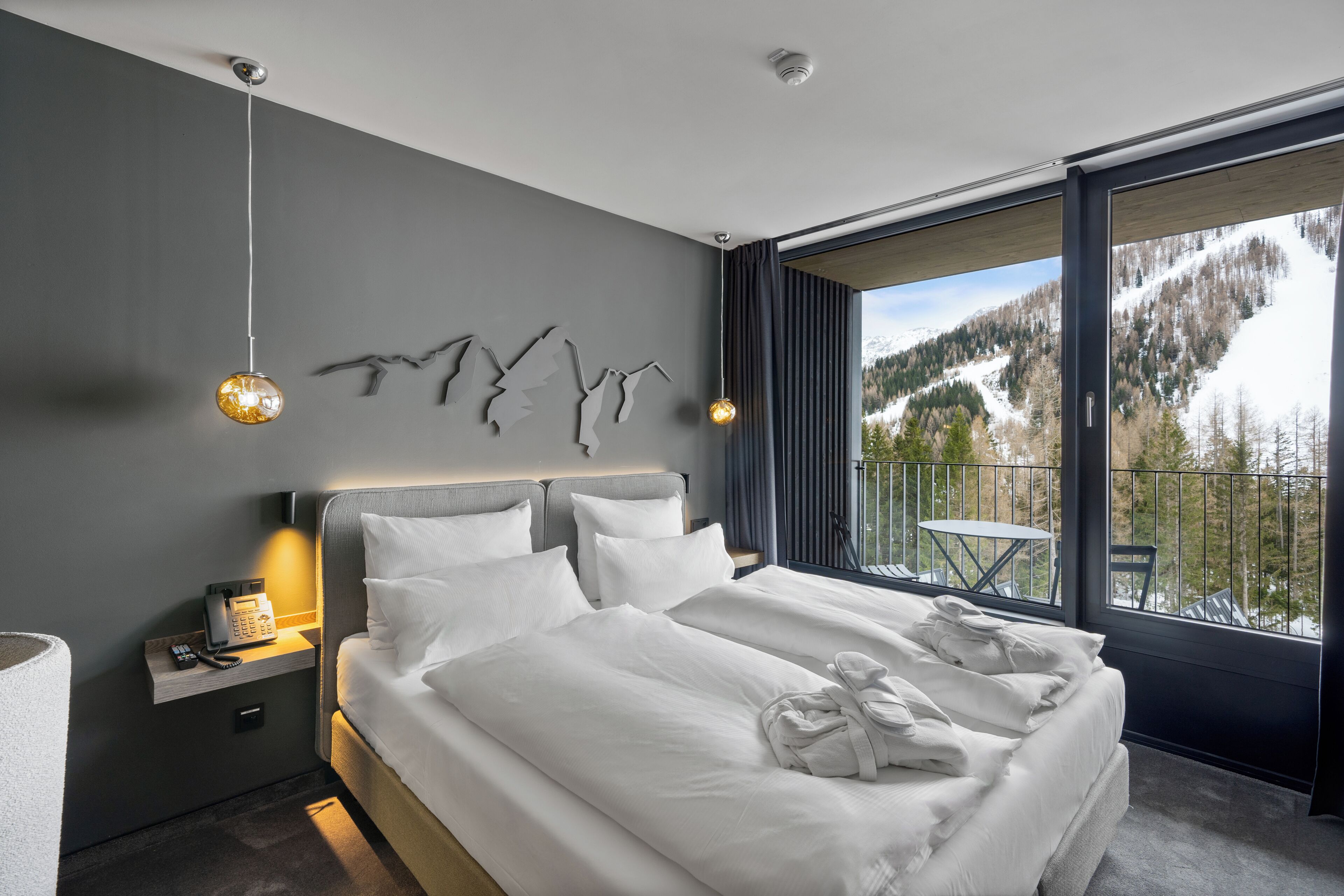 Ein modernes Hotelzimmer mit einem großen Bett, einem Nachttischtelefon, einem kleinen Tisch und einem Balkon mit Bergblick.