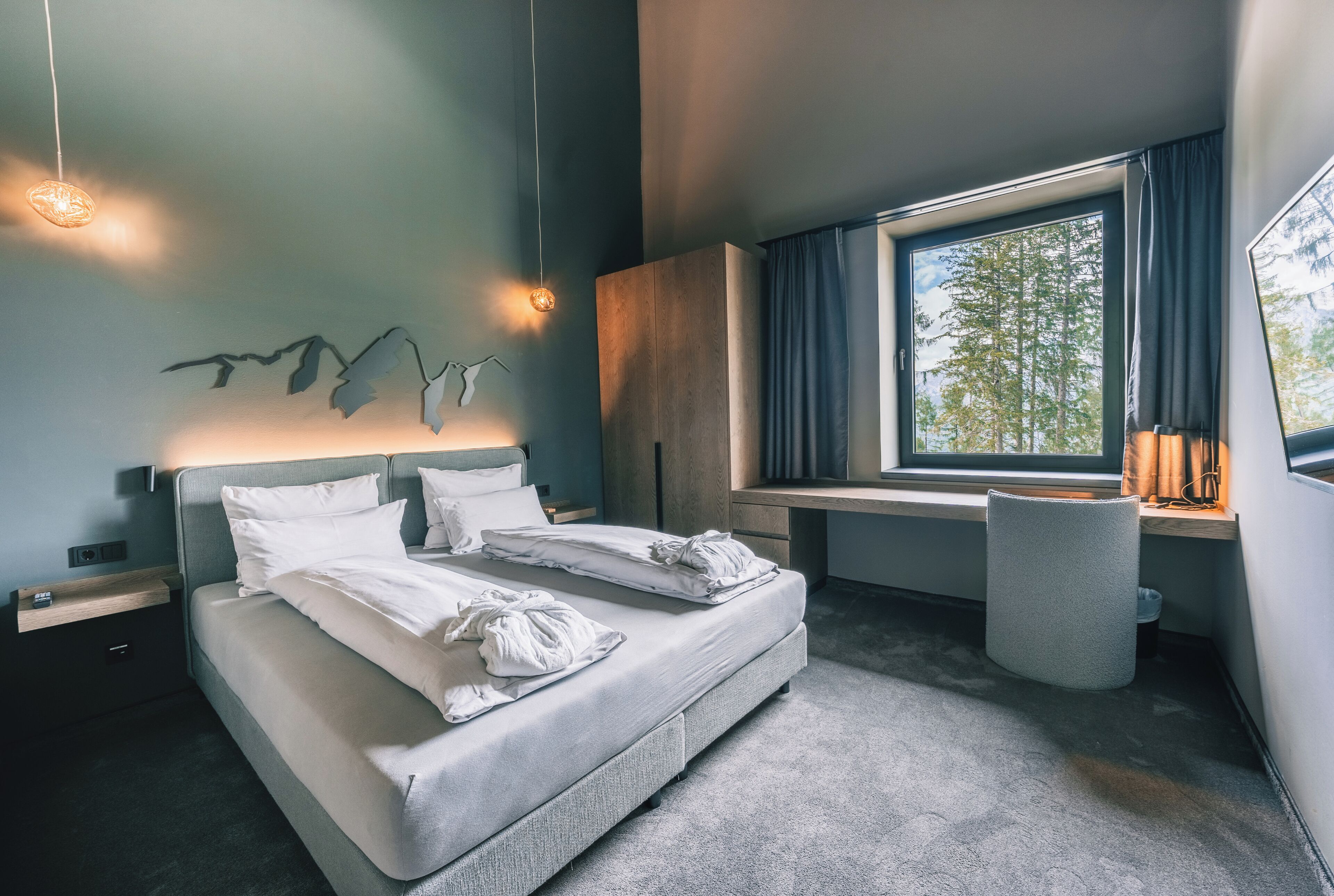 Un dormitorio moderno con cama, mesitas de noche, escritorio y ventana con vista a árboles.
