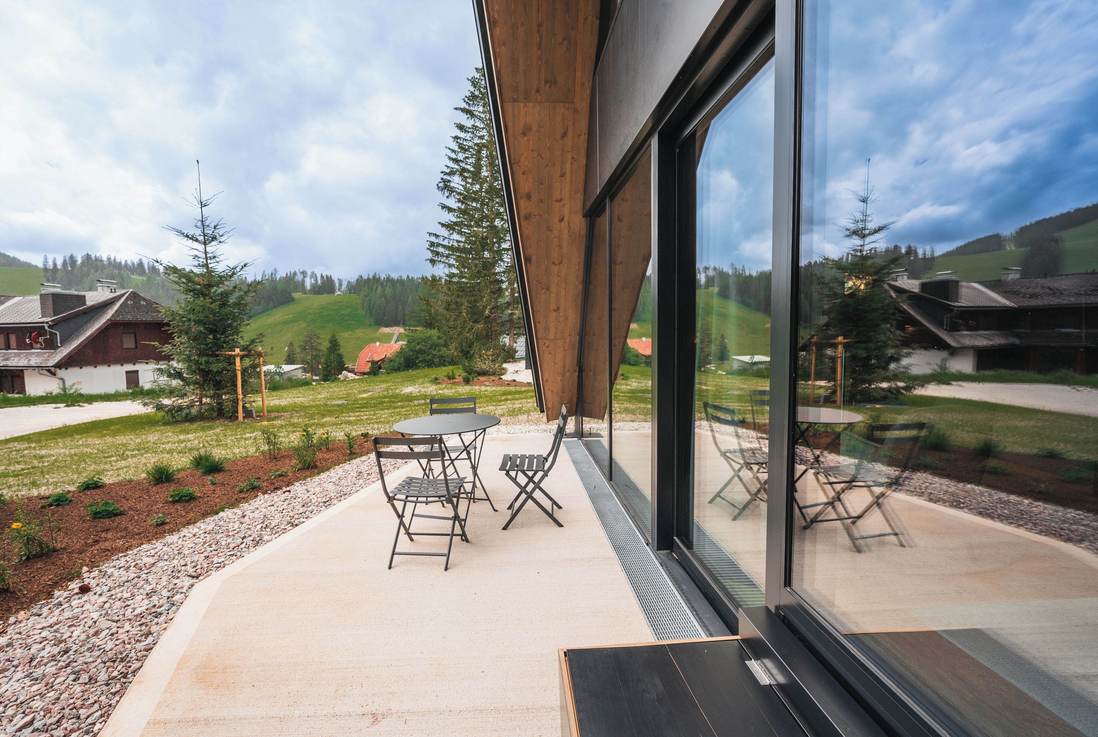 A modern building with large windows, outdoor seating, and a view of a green landscape.