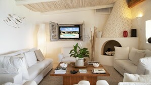 Una sala de estar con televisor, sofás blancos, una mesa de centro con libros y controles remotos, y una planta decorativa.