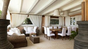 Un amplio espacio exterior para comer con mobiliario de mimbre y cojines blancos, rodeado de grandes ventanales con cortinas blancas.