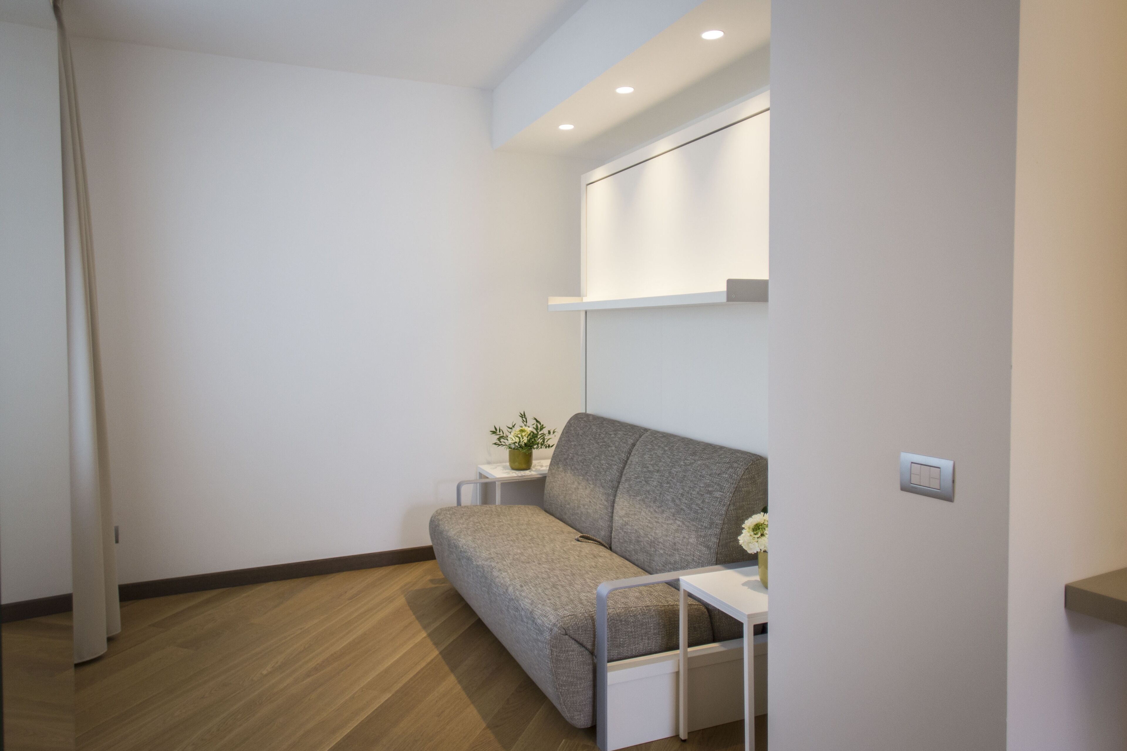 Une pièce moderne et minimaliste, avec un canapé gris, une petite table d’appoint blanche agrémentée d’une plante, et une étagère fixée au mur.
