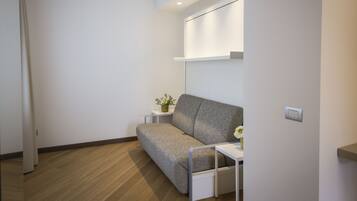 Una habitación moderna y minimalista con un sofá gris, una mesita auxiliar blanca con una planta y una estantería de pared.