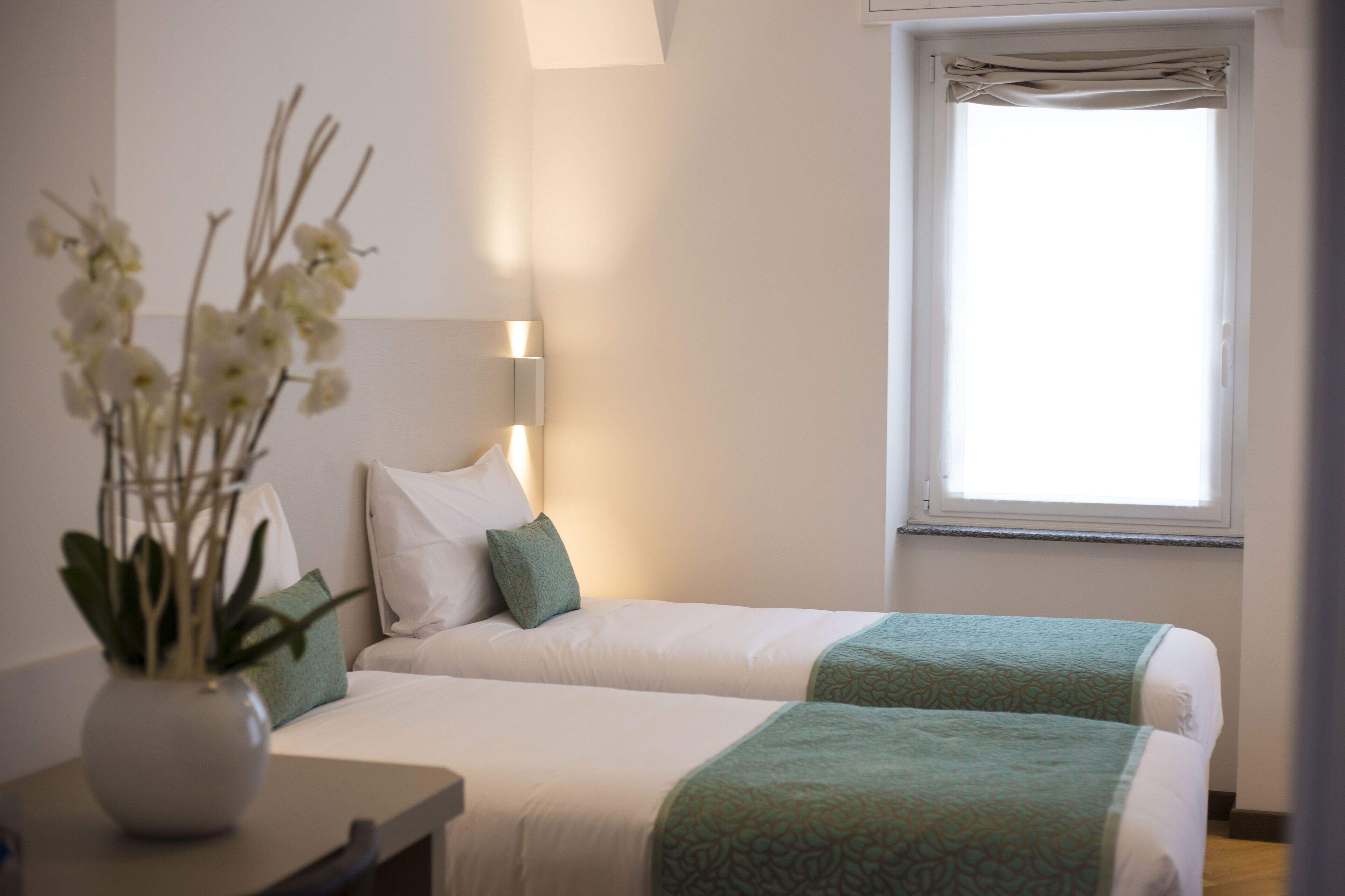 A hotel room with two beds, a bedside table, a vase with flowers, and a window with curtains.
