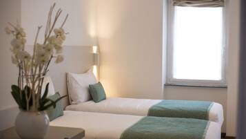 A hotel room with two beds, a bedside table, a vase with flowers, and a window with curtains.