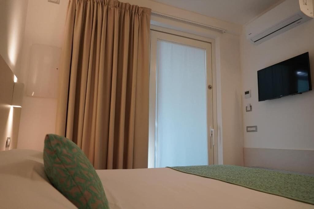 A hotel room with a bed, a television mounted on the wall, a window with curtains, and an air conditioning unit.