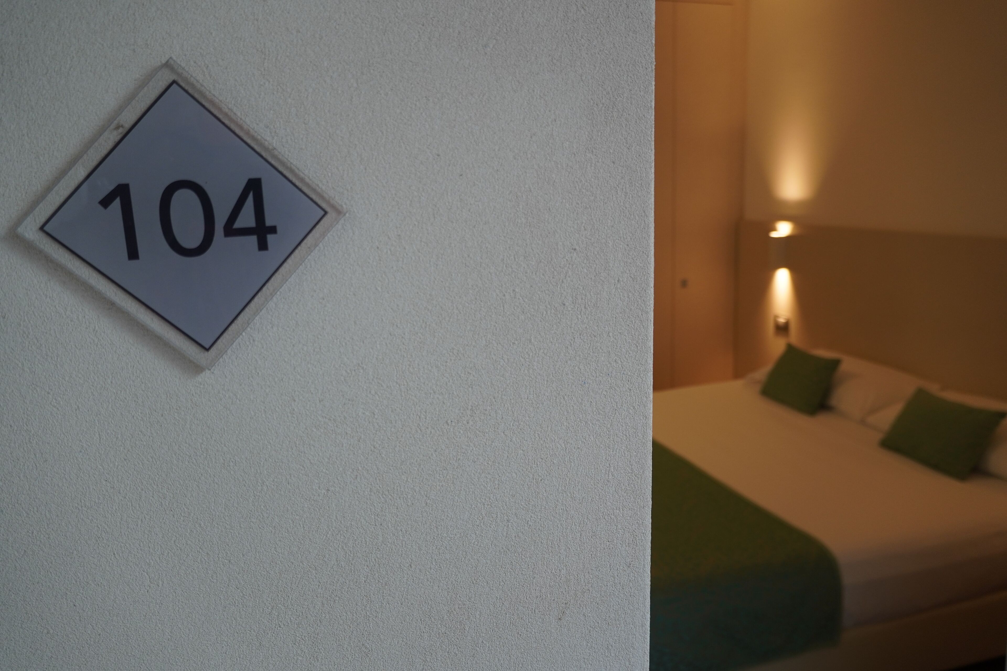 A room with a bed, a wall-mounted sign with the number 104, and a bedside lamp.