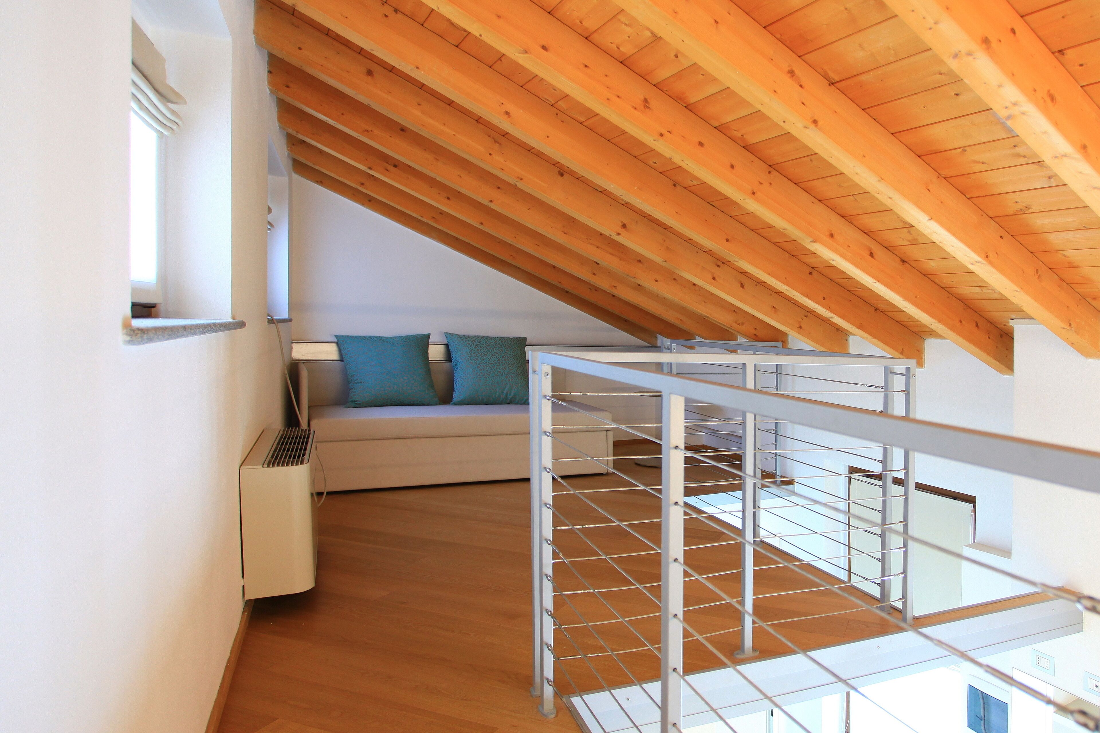 Ein modernes Schlafzimmer im Loftsstil mit schrägverlegtem Holzboden, einem weißen Sofa mit türkisfarbenen Kissen und einer Metalltreppe.