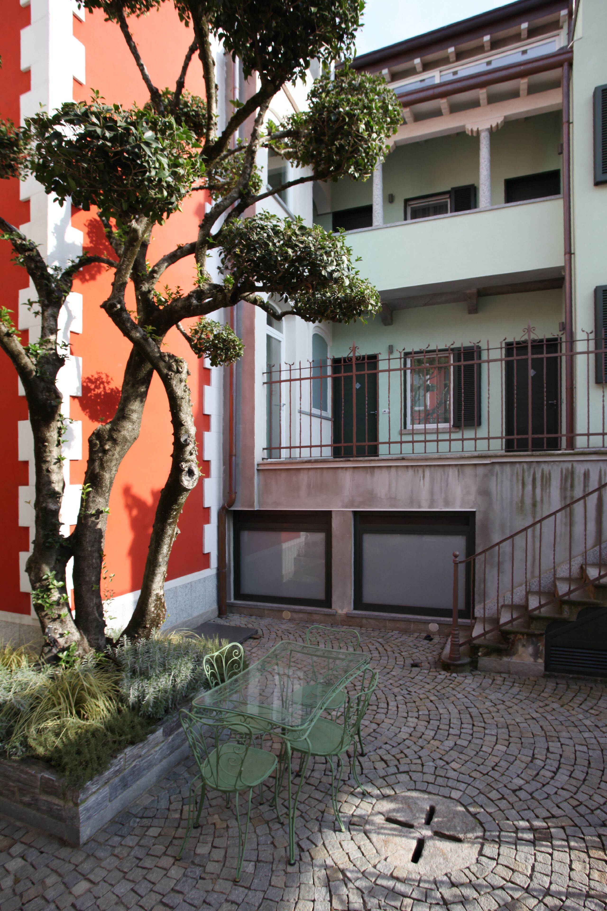 A courtyard with a tree, a glass table, and chairs. There is a building with a red and white facade in the background.